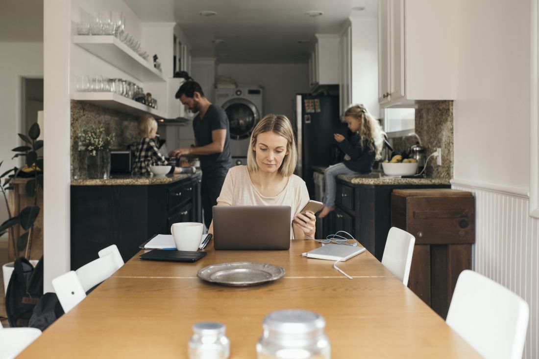 A woman sits at her kitchen table working on a laptop. A man and two children are in the background.