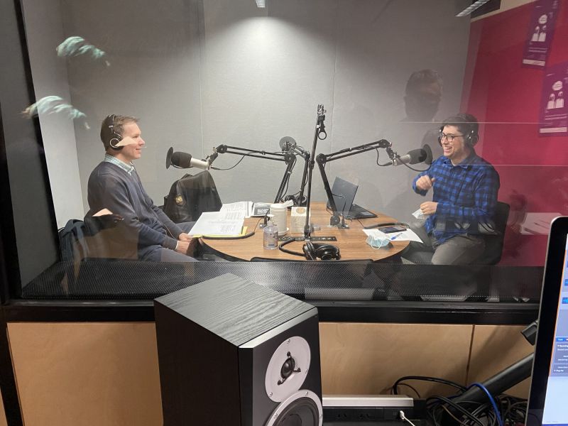 Outside the podcast studio, looking through a window to where where two people sit recording.