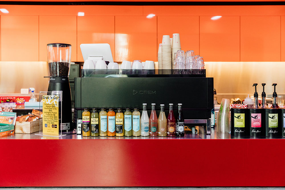 Photo of the red cafe counter, with juice and other drink bottles lined up on it. The coffee machine is behind the bottles.