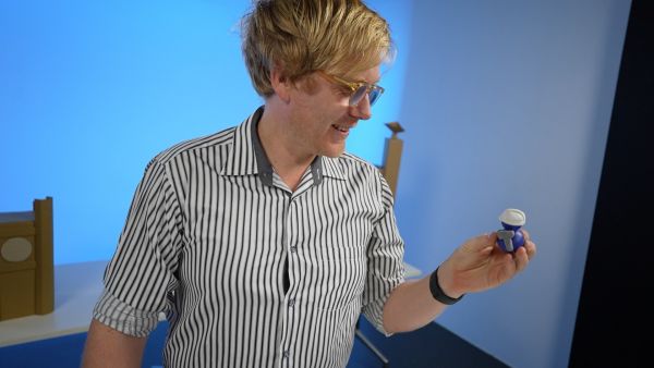 A man wearing a striped shirt stands in a blue-lit studio looking at a small blue clay model figure.