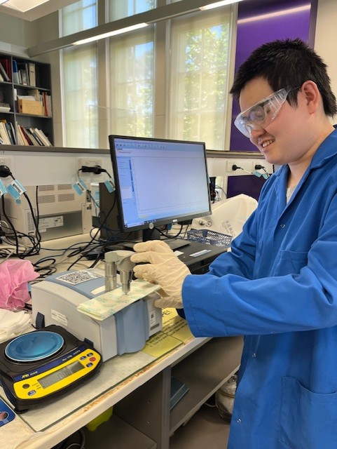 A dark haired man wearing safety goggles and a blue lab coat, working in the Healthcare Carbon Lab