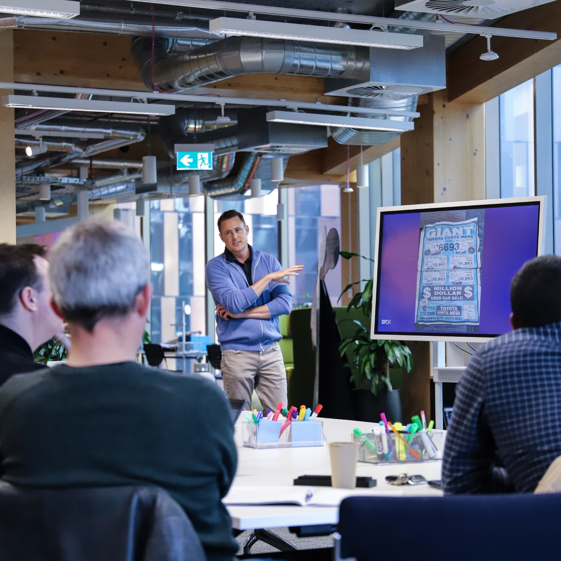 Spencer standing in front of a digital screen and speaking to a group sat around a table