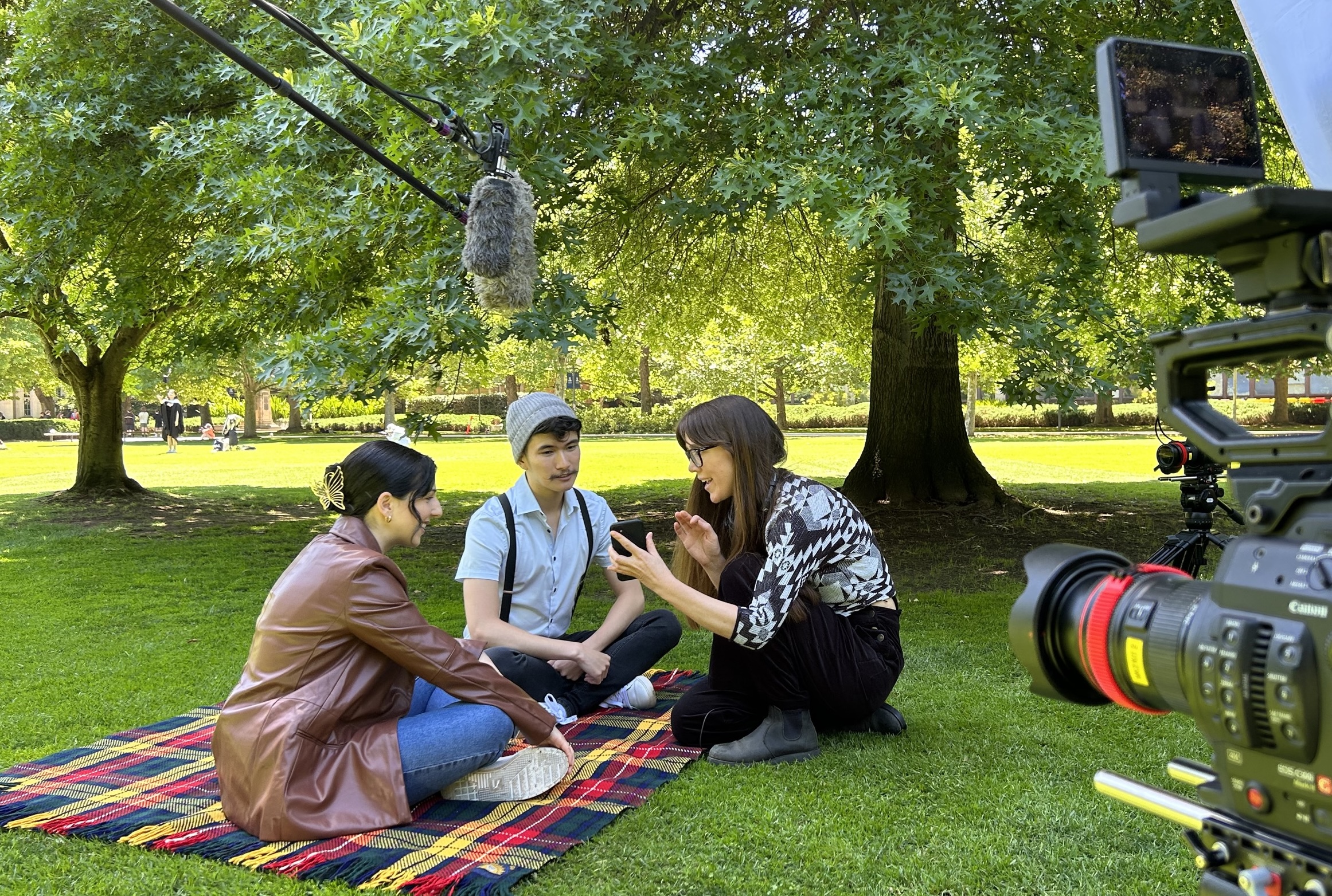 A film set filming two students sitting on a picnic blanket on a lawn on Parkville campus, under some trees - the director kneels down in front of them to give the students some advice about being on camera.