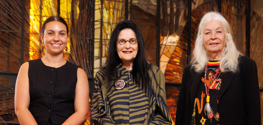 Three women stand together in front of a colorful stained glass window.