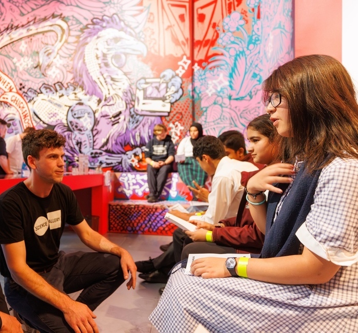 An individual speaking with several students in front of a colourful wall. 