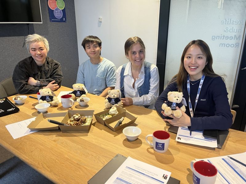 Four smiling students sit around a table with food and tea, holding small Barry the Bear plushies.
