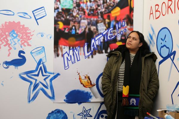 Amina Briggs stands in front of her colourful artwork wearing an green coat and an Aboriginal flag scarf.