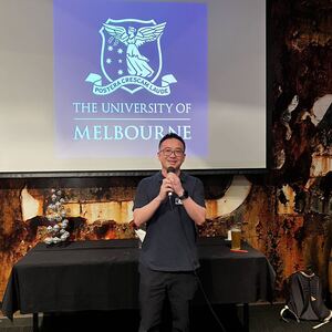 A man speaking at an alumni event. He is holding a microphone and the projection behind him is the University of Melbourne logo.
                                class=
