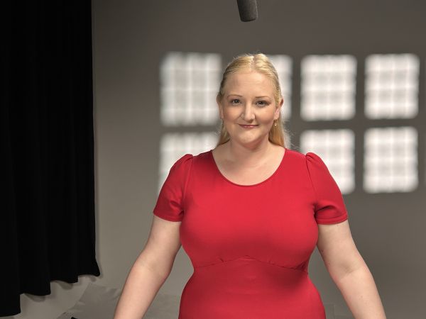 A woman with blonde hair and a bright red dress stands in front of a studio background with interesting lighting behind her.