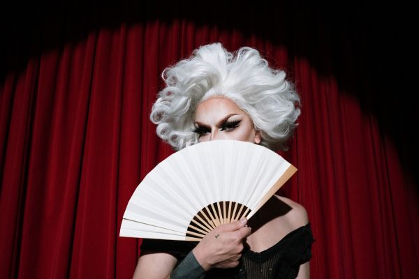 A photo of a drag queen with white hair in a studio with red drapes. Her face is behind a fan, with only her eyes showing. 