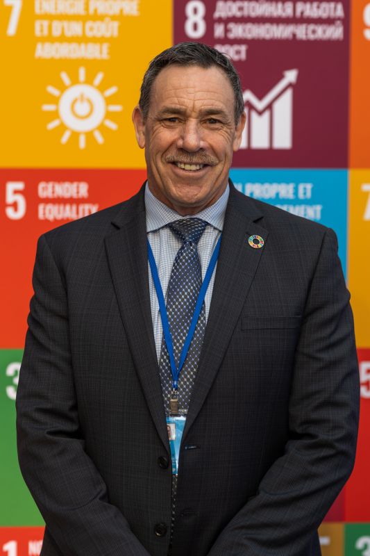 A dark haired man wearing a dark suit and standing in front of a large and colourful Sustainable Development Goals banner