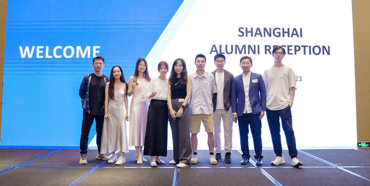 A group of nine alumni standing infront of a display that reads 'Shanghai Alumni Reception'.