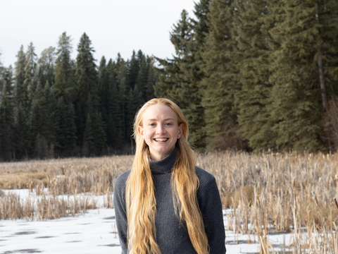 A lady with long blond hair stands surrounded by tall trees. 