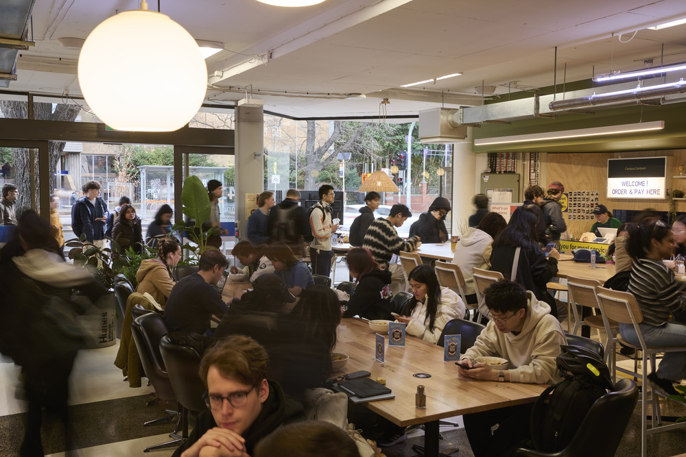 A crowd of people chatting and enjoying meals in a canteen setting.