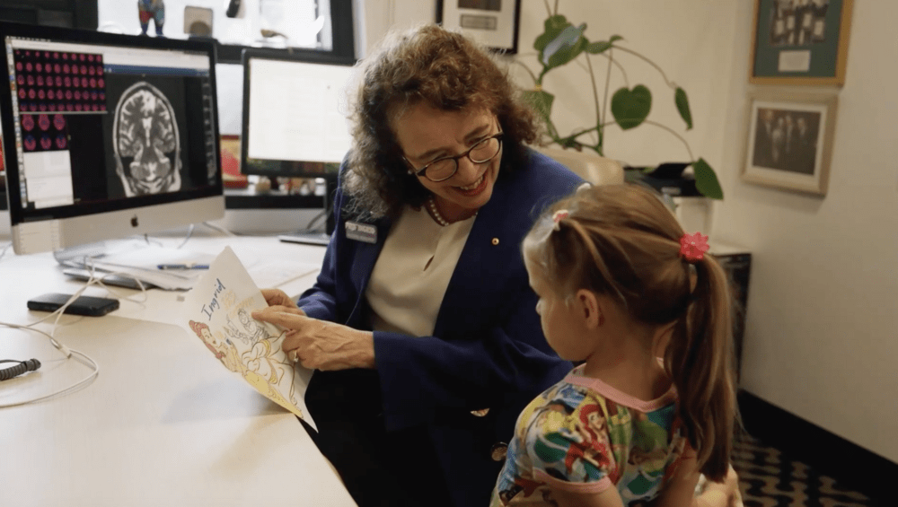 A woman and a young girl sit together at a desk, looking at a cartoon drawing.