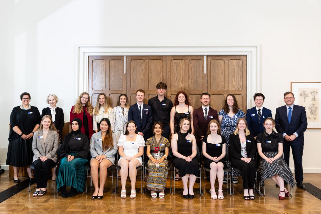Nine seated young women and nine standing young people, with two women at one end and one man at the other, are well dressed and smiling, stood in front of large timber doors in a white wall