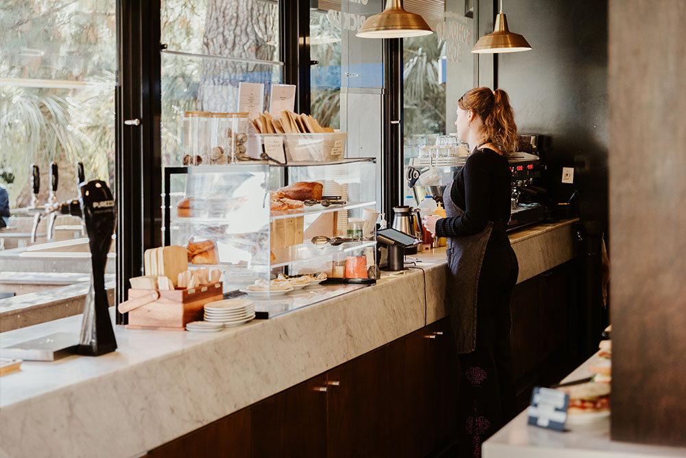 A woman standing behind a serving counter at Scholar & Co