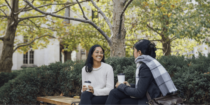 Two students seated on a bench surrounded by greenery.
