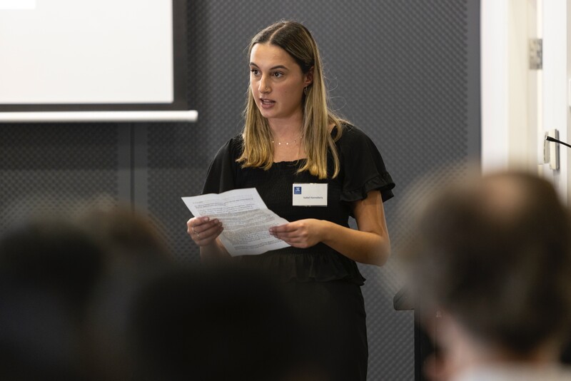 A female student is standing in front of a group of pepole and presenting in a room.