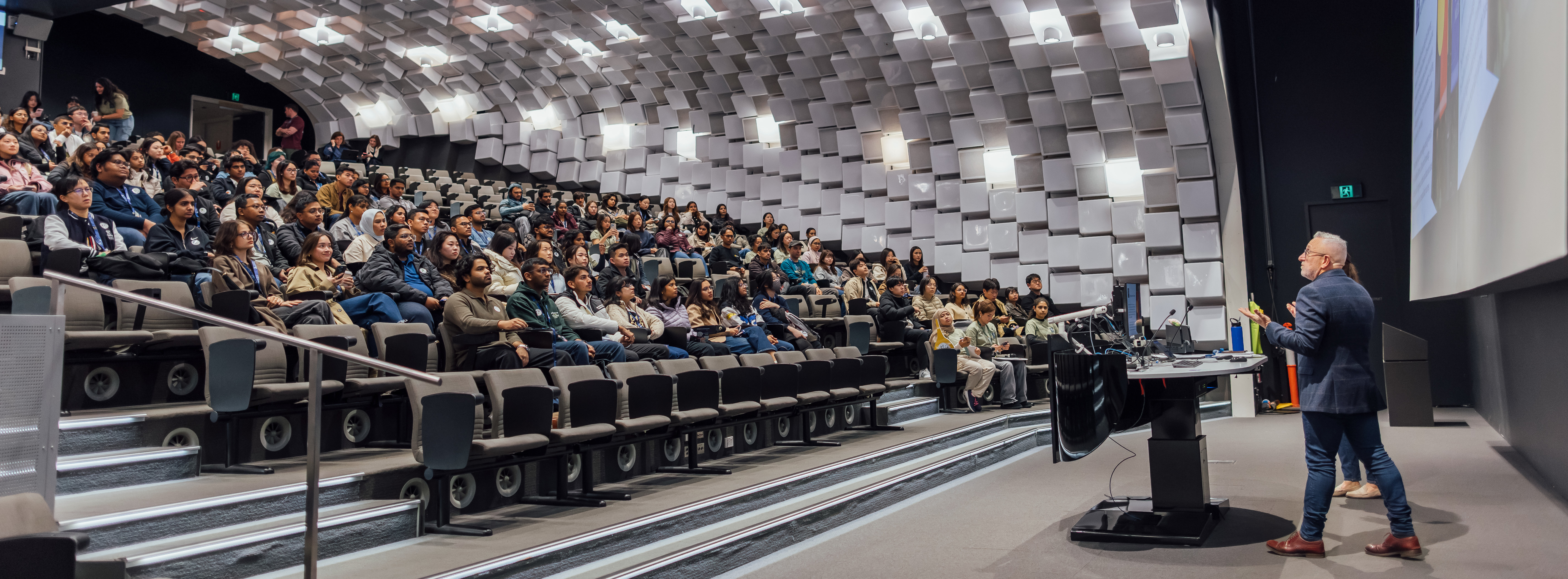 A speaker presenting in front of an audience in a lecture theatre.