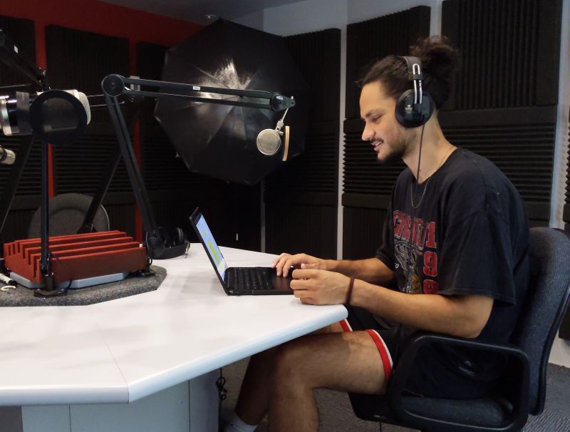 Photograph of a man wearing headphones and sitting at a desk in a soundproofed recording studio, working on a laptop. The setting includes a large microphone on an adjustable arm and acoustic foam panels on walls, indicating a podcast or audio recording environment.