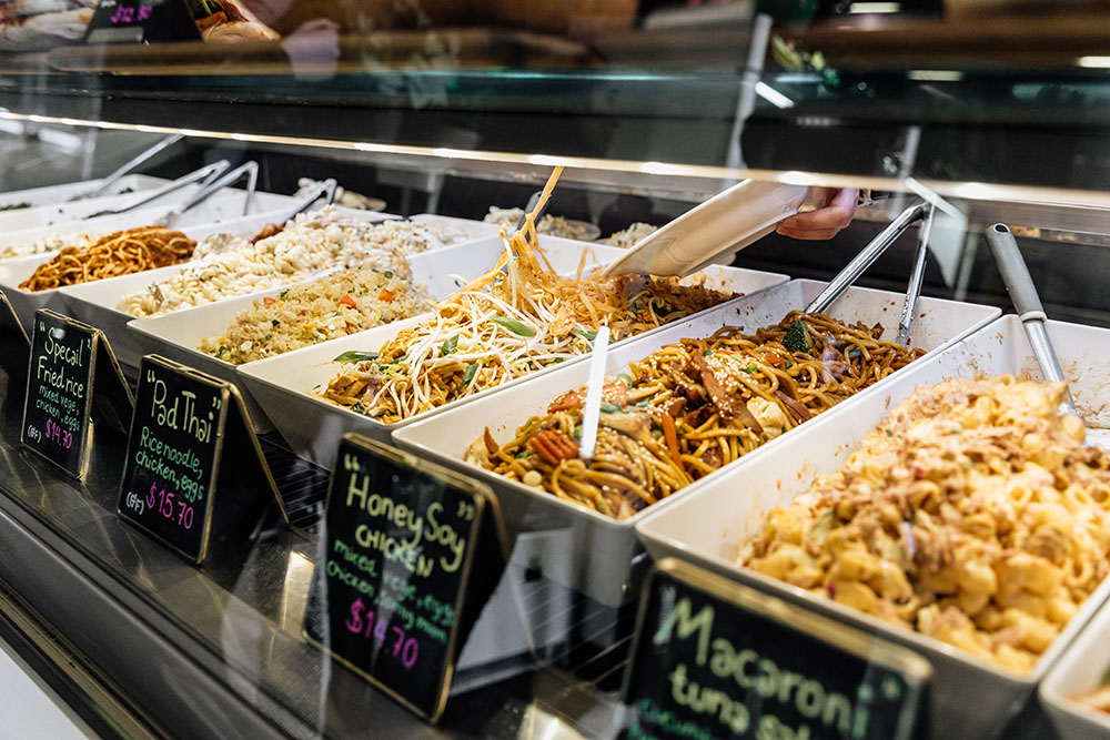 A row of bain-maries with food such as honey soy chicken noodles and Pad Thai in them