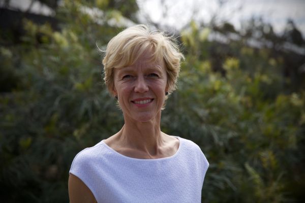 Woman with short blonde hair and a white top standing in front of shrubbery.