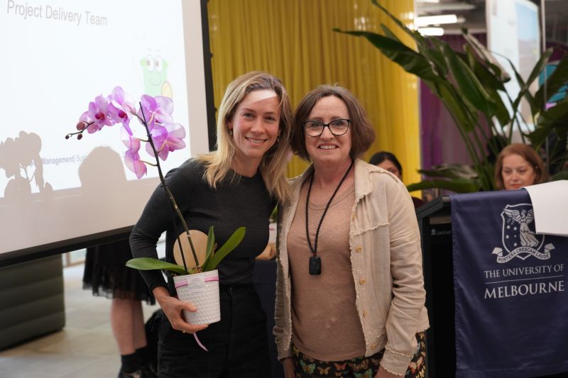 A blonde woman in a black top holds an orchid. She stands next to woman in a beige jacket