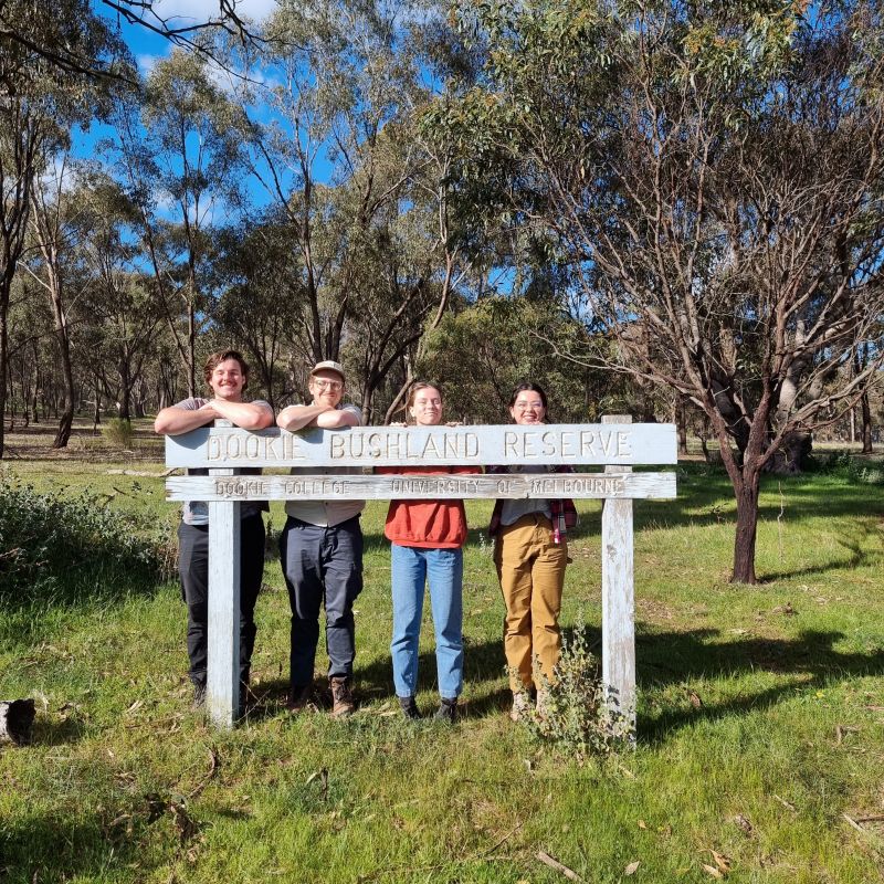 Four students standing behind a white wooden sign that says Dookie Bushland Reserve with trees behind them