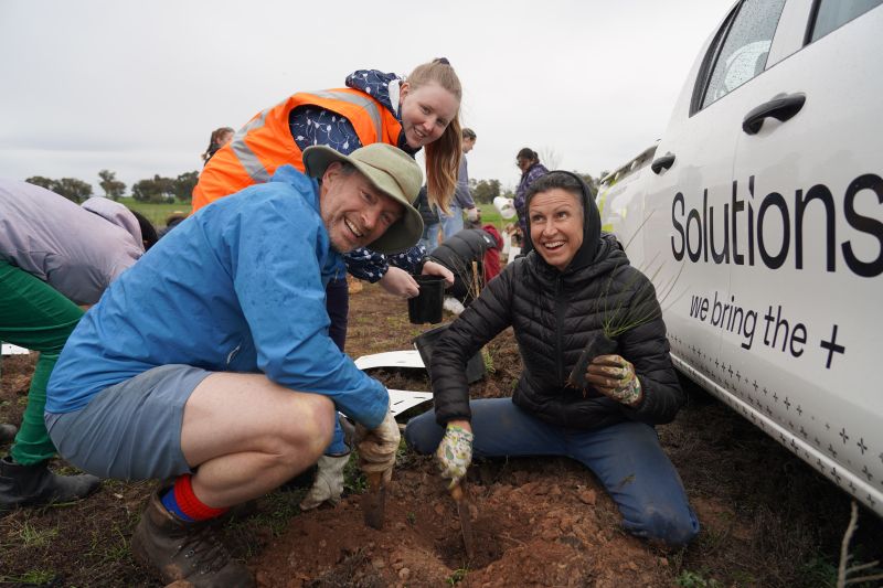 People planting small plants in soil beside a vehicle during an outdoor activity.