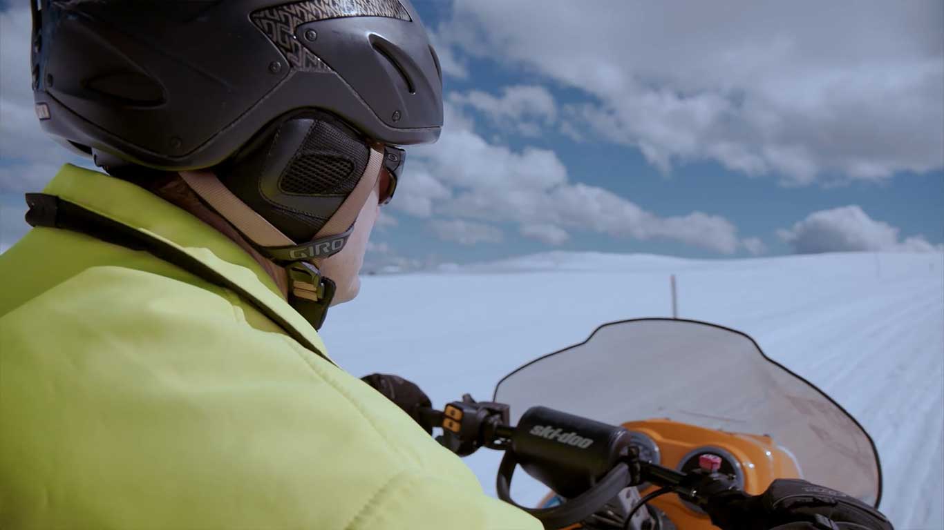 A man in high vis rides a snow scooter through the snow towards a blue, cloudy sky.