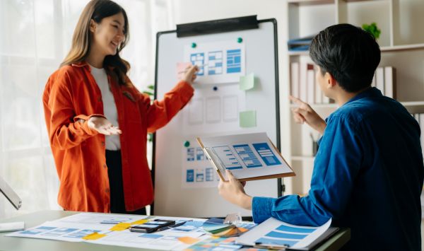 A woman and a man stand in front of a whiteboard poring over data research papers.