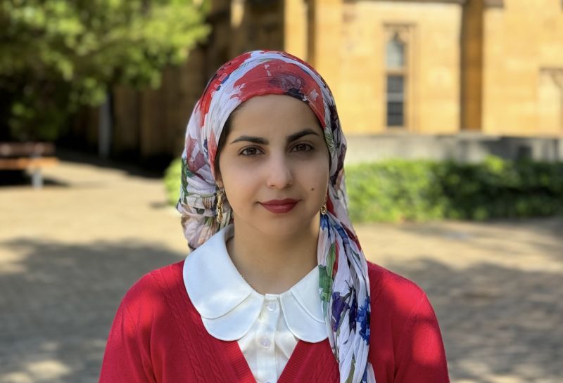 a smiling woman in a hijab and red cardigan and lipstick standing in front of trees and a sandstone building