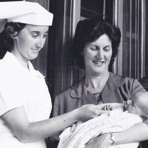 Black and white image of a woman cradling a baby with a nurse closeby.
                                class=