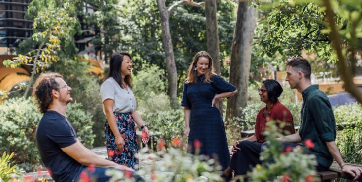 A group of five alumni conversing among trees and foliage with Arts West in the background.