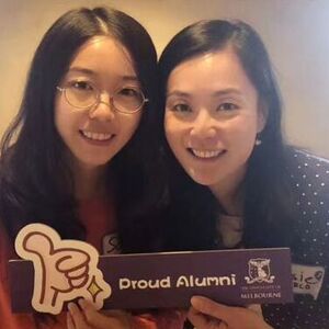 Two alumni volunteers smiling to camera, holding a sign that reads 'Proud Alumni'.
                                class=