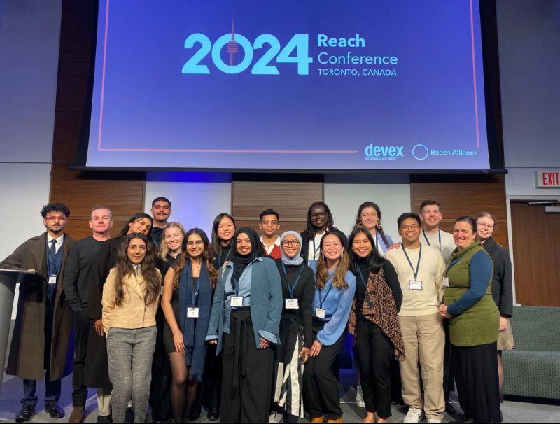 A group of around 20 staff and students underneath the blue Reach Conference banner