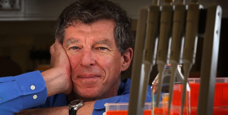 Ian Frazer smiling to camera while leaning on a desk with lab equipment in front of him.