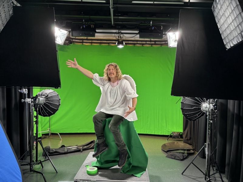 A man dressed as an angel sits in a film studio in front of a green screen.
