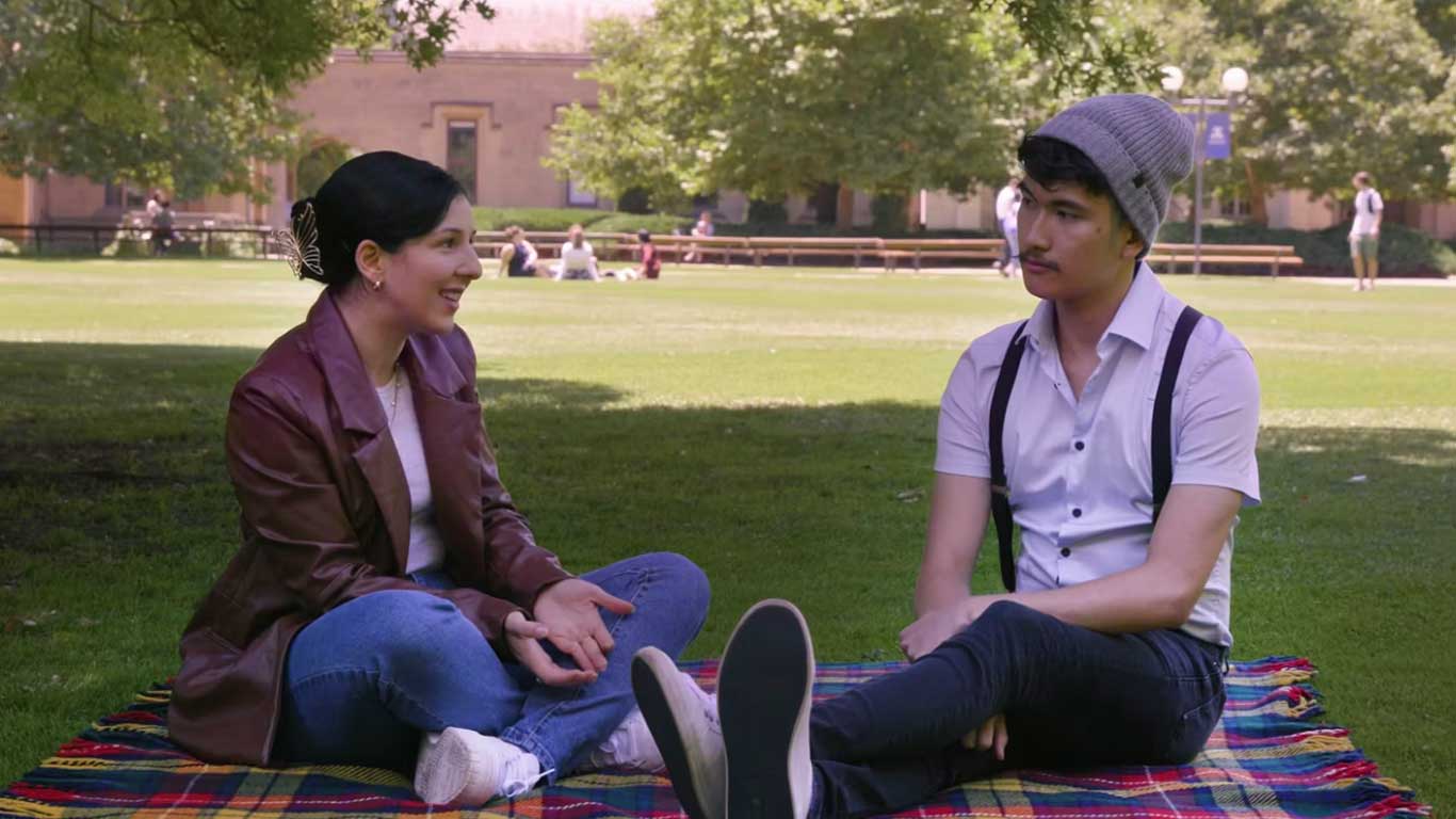 Two students sit on a picnic rug on south lawn talking.