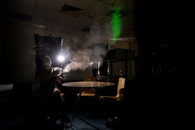 A man sits at a table in a dark room, pushing air out of a plastic bottle. A bright light shows how the air travels 