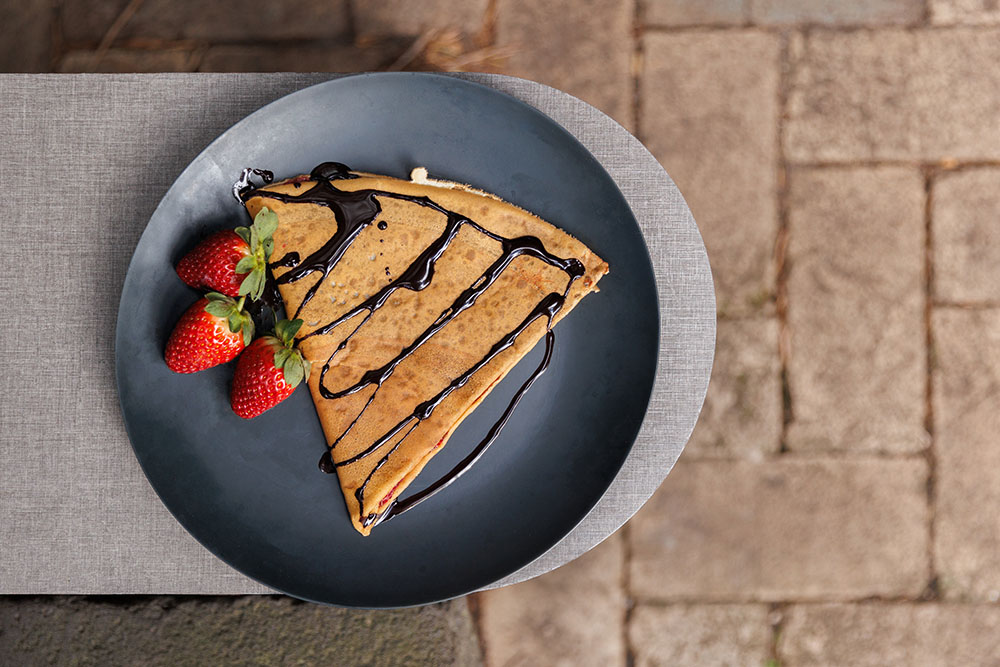 Looking down at a plate with a sweet crepe on it, drizzled with chocolate sauce and decorated with strawberries