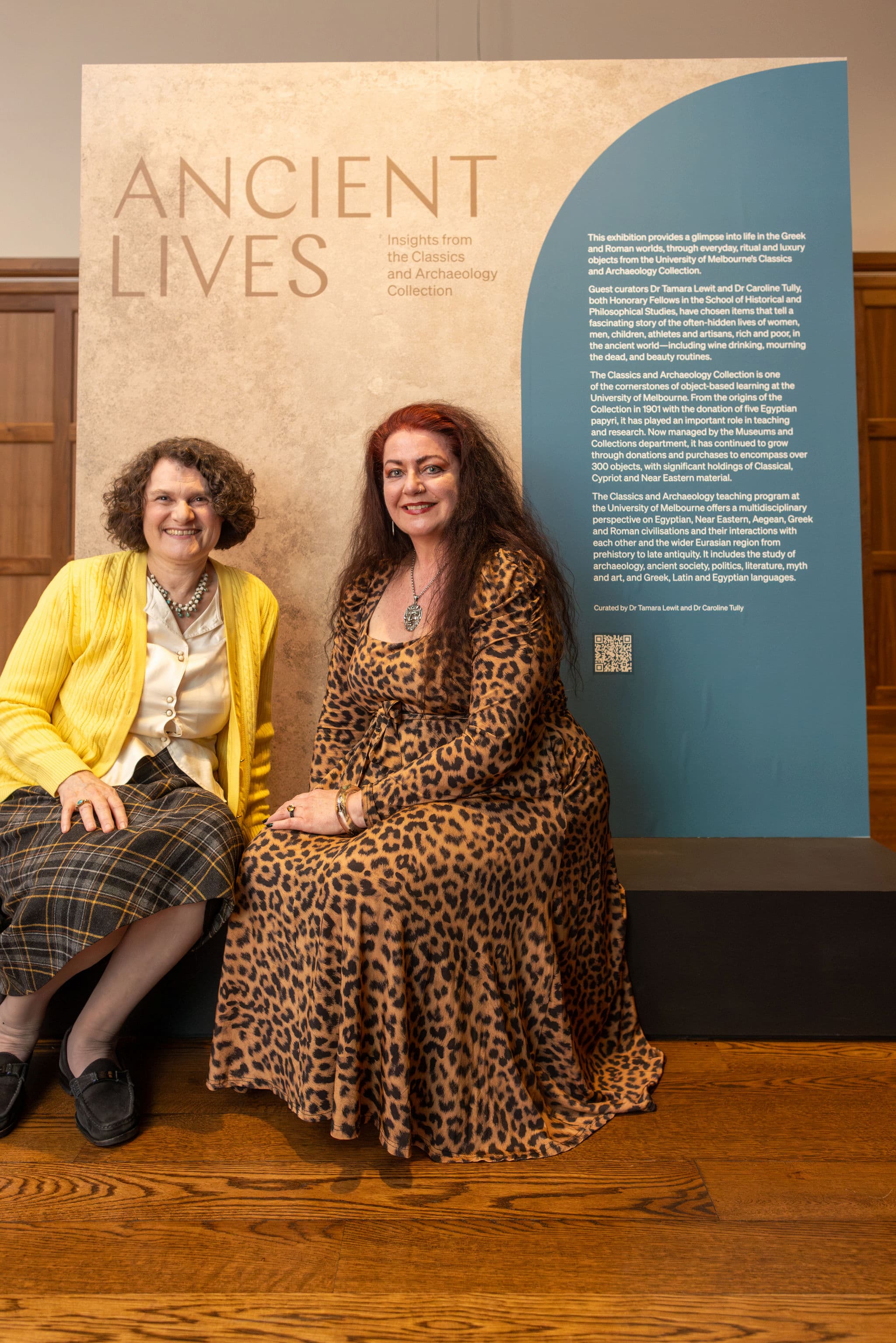 Two women sit in front of a sign for Ancient Lives