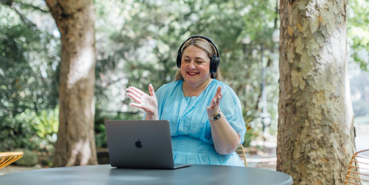 A woman sitting at a table wearing a headset and smiling during a video call on her laptop.