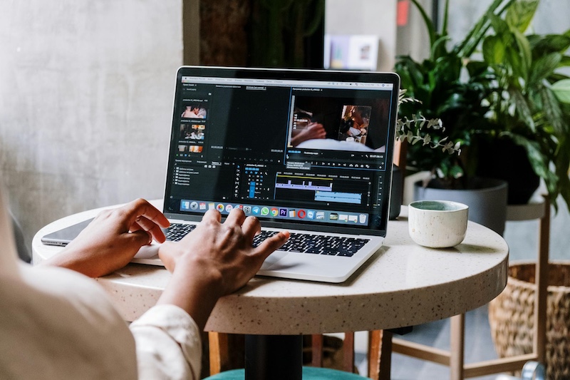 A woman is at her laptop at home working on a video editing timeline in video editing software. We can't see her face, just her back and arms.