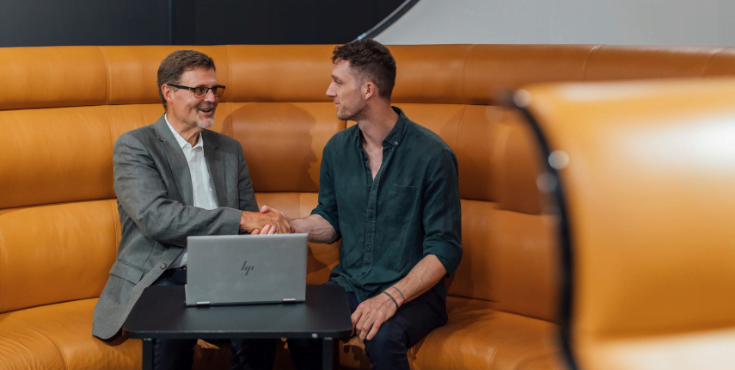 An older mentor and younger mentee sitting together, smiling and shaking hands.