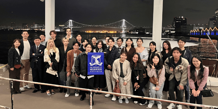 A large group of Japanese alumni posing for a picture together. They're holding a University of Melbourne flag in front of an expansive river.