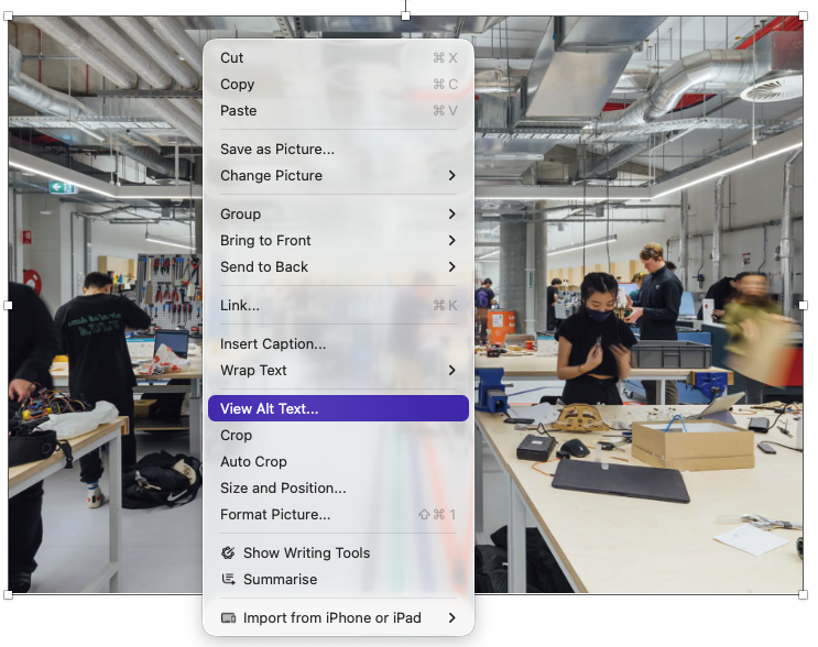 The right-click menu for images, containing the View Alt Text item Photograph of a modern workshop or makerspace with multiple people working at large tables equipped with tools, electronic components, and materials. The space features exposed ductwork, bright lighting, and coloured floor lines guiding pathways, highlighting an organised environment for collaborative technical or creative projects.