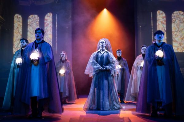 An actor in a robe and headpiece stands in the middle of six other actors in cloaks, carrying glowing orbs as part of The Magic Flute opera. 