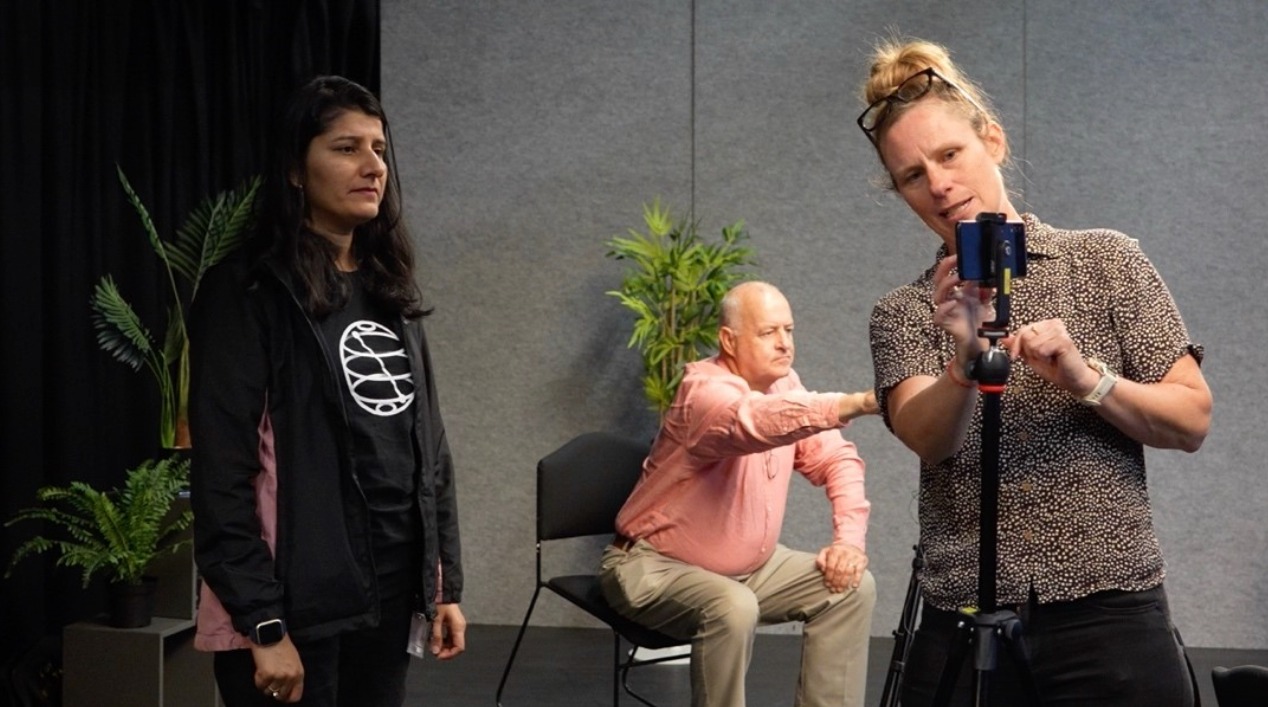 There are three people in a film studio. One woman stands in front of a device on a stand looking at camera. Another woman is adjusting the camera and showing her how to use it. Behind them, a man sits on a chair adjusting his own camera on a stand.